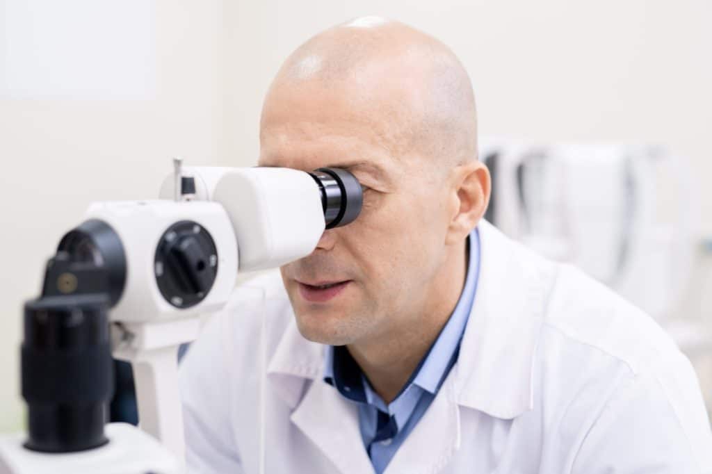 Professional ophthalmologist looking through medical equiment by workplace